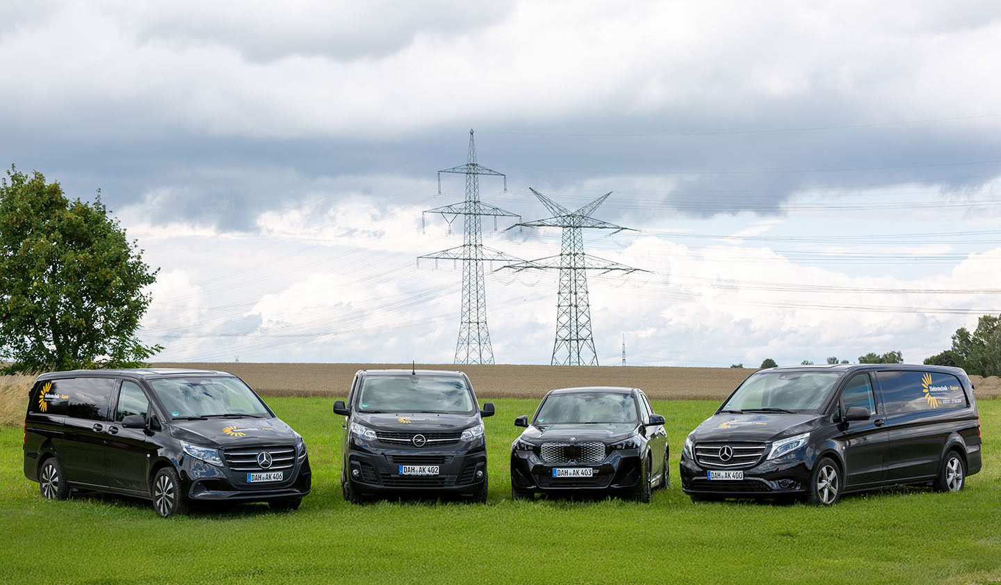 Vier schwarze Firmenfahrzeuge stehen auf einer Wiese vor einem Acker mit Hochspannungsmasten im Hintergrund.