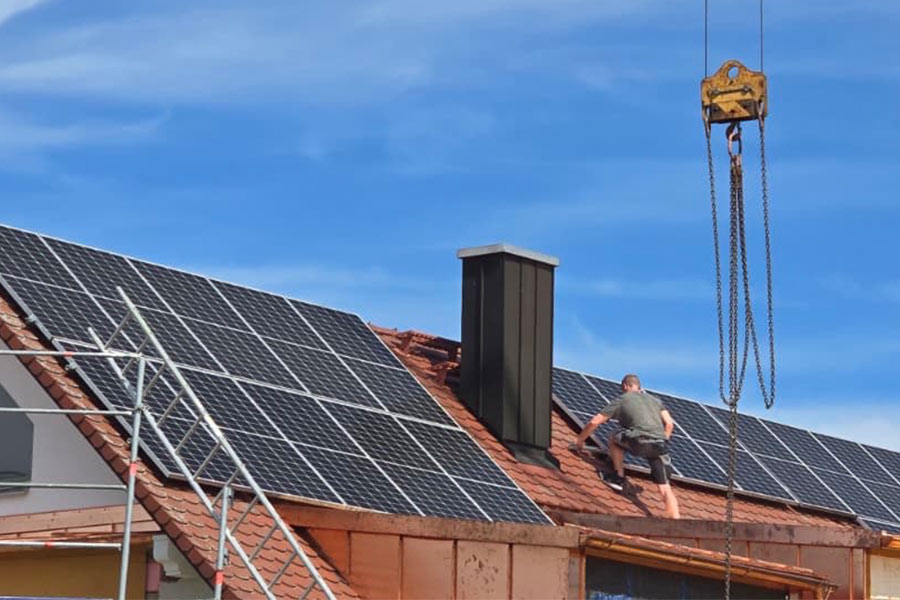 Ein Handwerker arbeitet auf einem Dach mit großen Solarmodulen, daneben ist ein Baukran sichtbar.