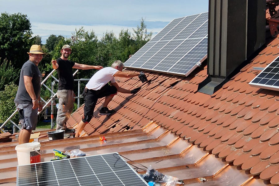 Drei Handwerker installieren Solarmodule auf einem Ziegeldach bei sonnigem Wetter.
