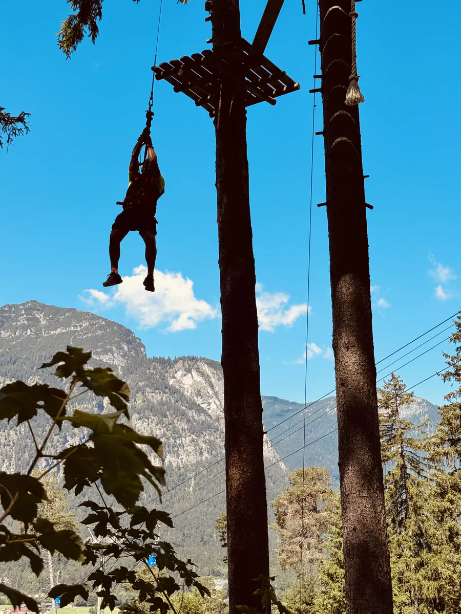Kollege hängt kopfüber gesichert an einem Klettergerüst vor Bergkulisse